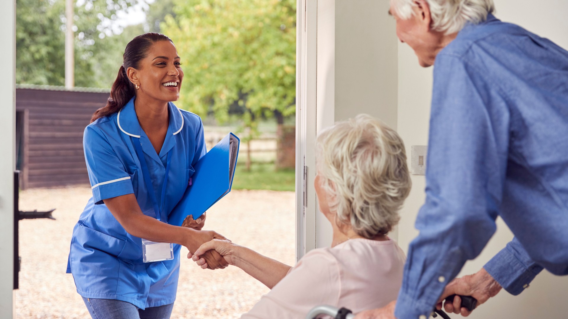 Senior Couple With Woman In Wheelchair Greeting Nurse Or Care Worker Making Home Visit At Door